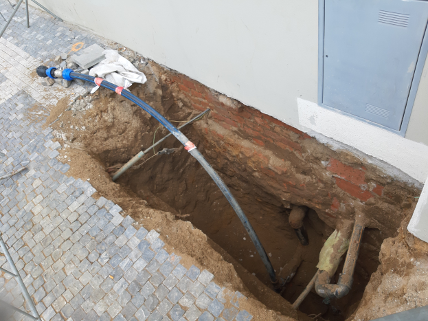 Ein großes Loch im Boden neben einem Gebäude, mit verstreuten Rohren und Gegenständen, einem Metallobjekt links und einem Metallkasten an der rechten Wand, wahrscheinlich eine laufende Kanalarbeitsdienstleistung.