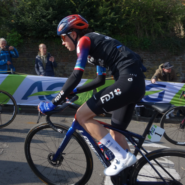 Eine professionelle Radsportlerin, Katie Archibald, fährt mit Helm, Brille, Handschuhen und Schuhen ein Fahrrad auf einer Straße neben einer Menge Menschen. Im Hintergrund gibt es eine Fahne mit Text, eine Wand, Bäume und ein Gebäude.