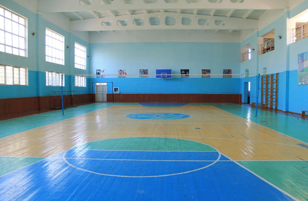 Große Turnhalle mit einem blauen und weißen Basketballfeld, Netz, Pfosten, einer Tür, Fenstern, Wandplakaten, einem Geländer und einem Dach.