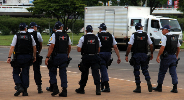 Eine Gruppe von Beamten in Uniform, einschließlich Mützen, Hemden, Schutzwesten, Uhren, Hosen und Schuhen, geht auf einer Straße. Rechts daneben steht ein Lastwagen in der Nähe eines Zaunes, mit Gebäuden, Bäumen, Pflanzen, Pfählen und Schildern im Hintergrund. Eine Brücke ist oben rechts zu sehen.