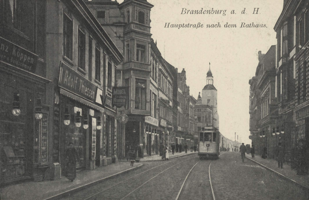 Ein Schwarz-Weiß-Foto einer Stadtstraße in Brandenburg, Deutschland, das eine Straßenbahn auf Schienen, Menschen auf dem Bürgersteig, Gebäude mit Fenstern, Schilder und Straßenlaternen unter einem bewölkten Himmel zeigt.