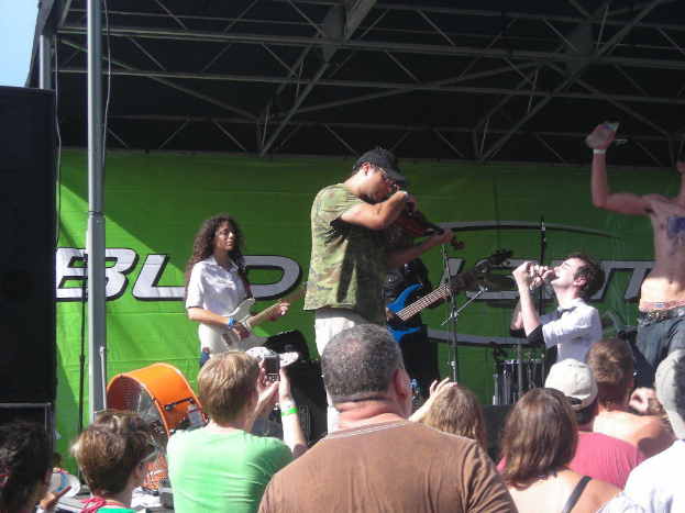 Eine Gruppe von Musikern, die verschiedene Instrumente auf einer Bühne spielen, während links ein großes schwarzes Lautsprechers steht und das Publikum vorne steht und die Performance genießt.