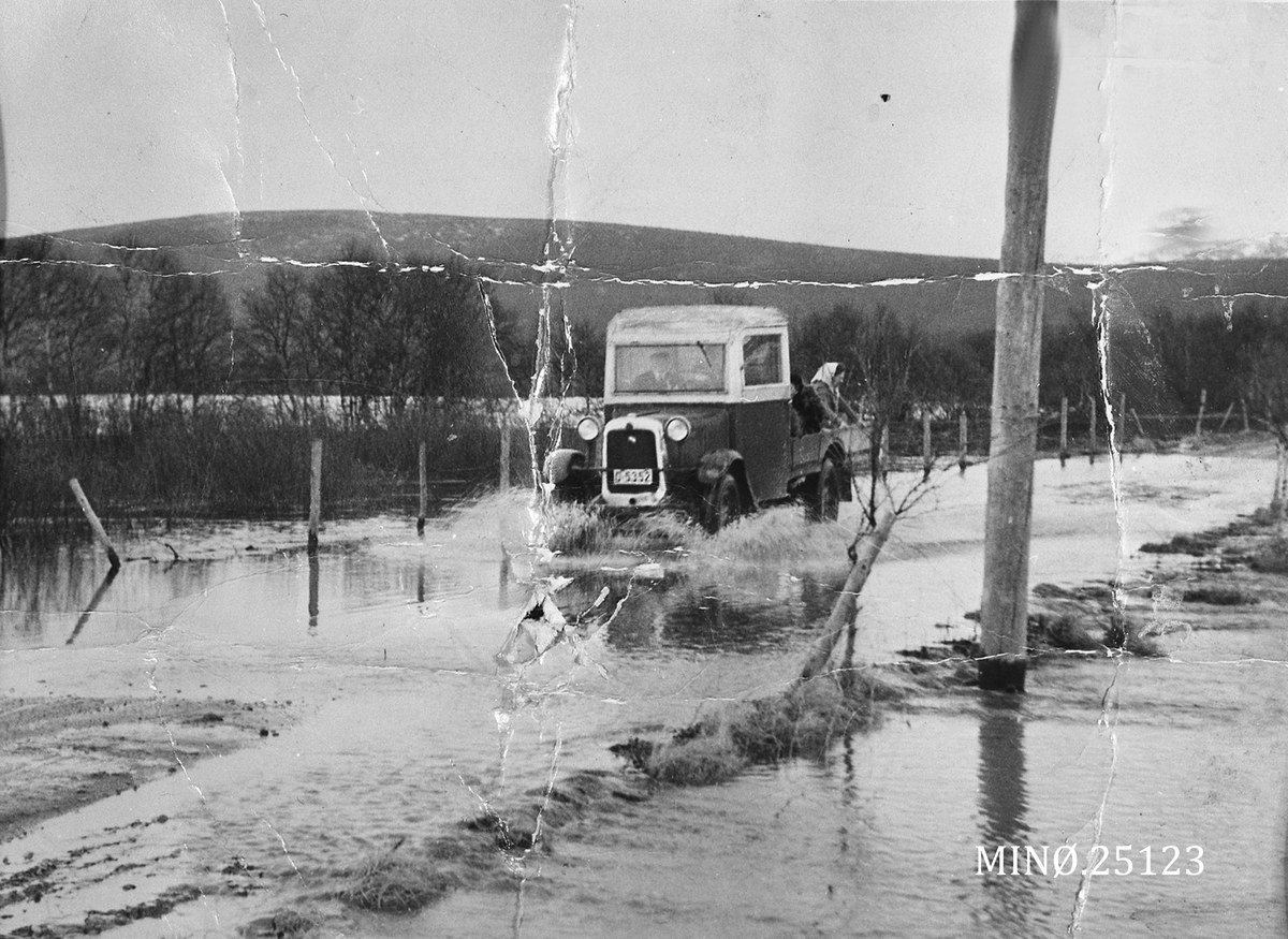 Ein alter Laster fährt durch eine überflutete Straße mit Wasser drumherum, Polen, Bäumen und einem Hügel im Hintergrund unter einem sichtbaren Himmel, mit einem Wasserzeichen in der rechten unteren Ecke.