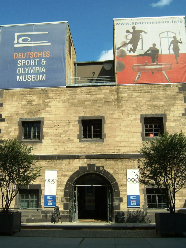 Außenansicht des Deutschen Sport & Olympiamuseums in Berlin mit Fenstern, einer Tür, umgeben von Bäumen und Bannern mit Text und Bildern unter einem bewölkten Himmel.