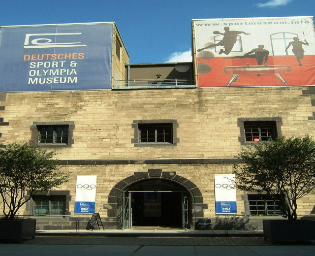 Außenansicht des Deutschen Sport & Olympiamuseums in Berlin mit Fenstern, einer Tür, umgeben von Bäumen und Bannern mit Text und Bildern unter einem bewölkten Himmel.