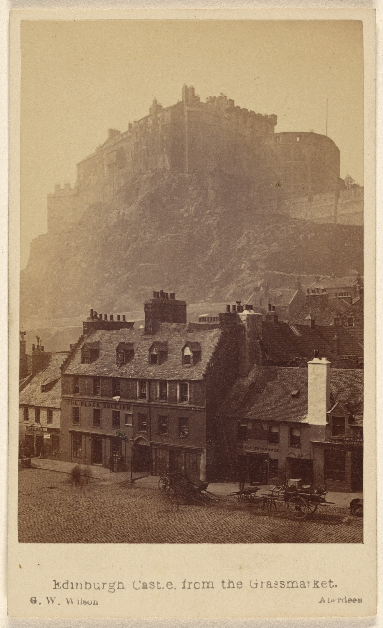 Alte Fotografie von Edinburgh Castle aus der Grassmarket-Sicht, mit Gebäuden und Karren im Vordergrund, einem Hügel im Hintergrund und Text am unteren Rand.