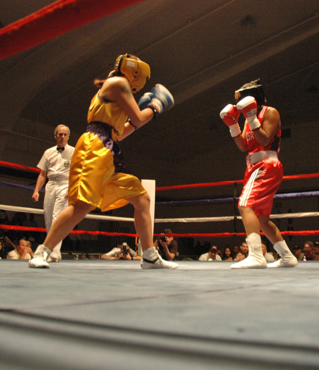 Zwei Boxer kämpfen im Ring mit einem Schiedsrichter und Zuschauern, einige halten Kameras, unter einer sichtbaren Decke.