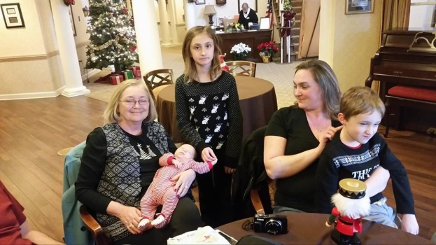 Ein Raum mit einer Wand, die ein Photo-Frame, einen Weihnachtsbaum, eine Lampe und Blumenpflanzen zeigt. Im Vordergrund gibt es Treppen und einige Menschen, die an Tischen sitzen, mit einem Spielzeug und einer Kamera auf einem der Tische.