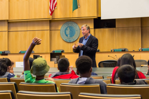 Ein Mann in einem schwarzen Blazer, blauer Hemd und schwarzen Hosen steht vor einer Klasse aufmerksamer Kinder, h├Ąlt ein Mikrofon in der Hand, mit einer h├Âlzernen Wand, Logo, zwei Flaggen und einem wei├čem Bildschirm dahinter.
