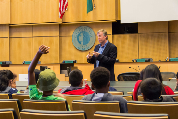 Ein Mann in einem schwarzen Blazer, blauer Hemd und schwarzen Hosen steht vor einer Klasse aufmerksamer Kinder, h├Ąlt ein Mikrofon in der Hand, mit einer h├Âlzernen Wand, Logo, zwei Flaggen und einem wei├čem Bildschirm dahinter.