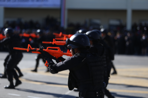 Eine Gruppe von Polizisten in Einsatzausrüstung, mit Helmen und Waffen bewaffnet, marschiert eine Straße entlang mit einem Gebäude und einer Fahne mit Text im Hintergrund.
