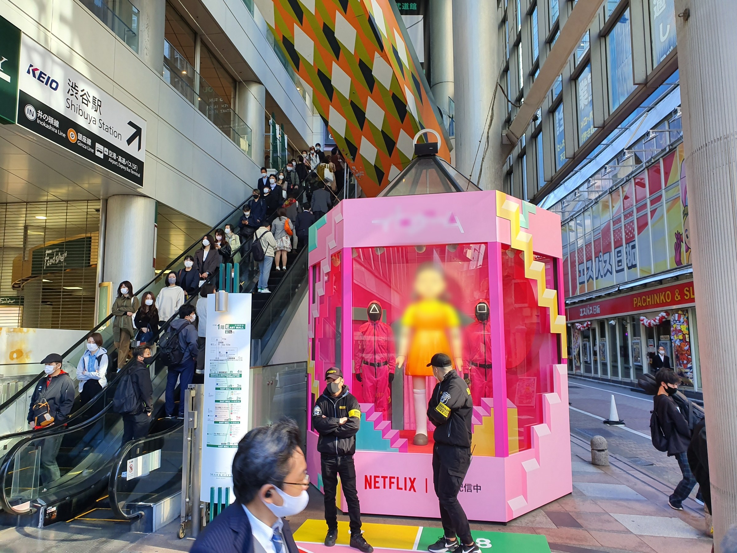 Eine Gruppe von Menschen steht vor einem Gebäude mit Glaswänden, Säulen und Texttafeln, einige tragen Masken, während eine Rolltreppe mit Menschen zu sehen ist. In der Mitte steht ein pinker Kasten mit der Aufschrift "Netflix's new Netflix booth in Tokyo", in dem Puppen zu sehen sind.