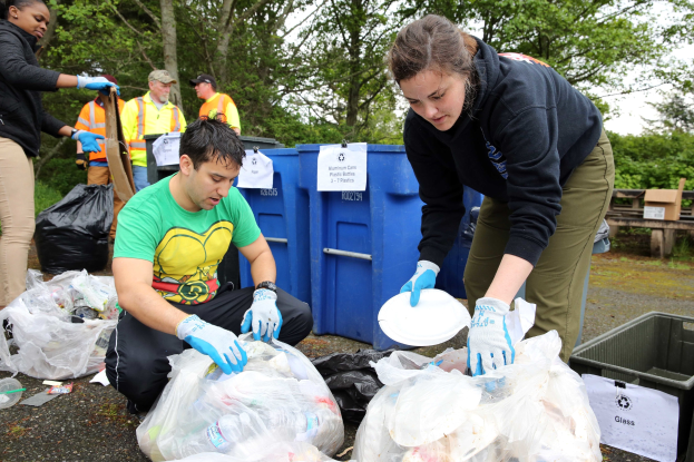 Eine Gruppe von Menschen, die Müll in einem Park sammeln, mit einem Mann und einer Frau in der Mitte, die Handschuhe tragen und Schilder halten, umgeben von Plastikabfällen, Flaschen und anderen Gegenständen auf dem Boden, ein Müllcontainer und eine Holzbank rechts und Bäume mit einem klaren blauen Himmel im Hintergrund.
