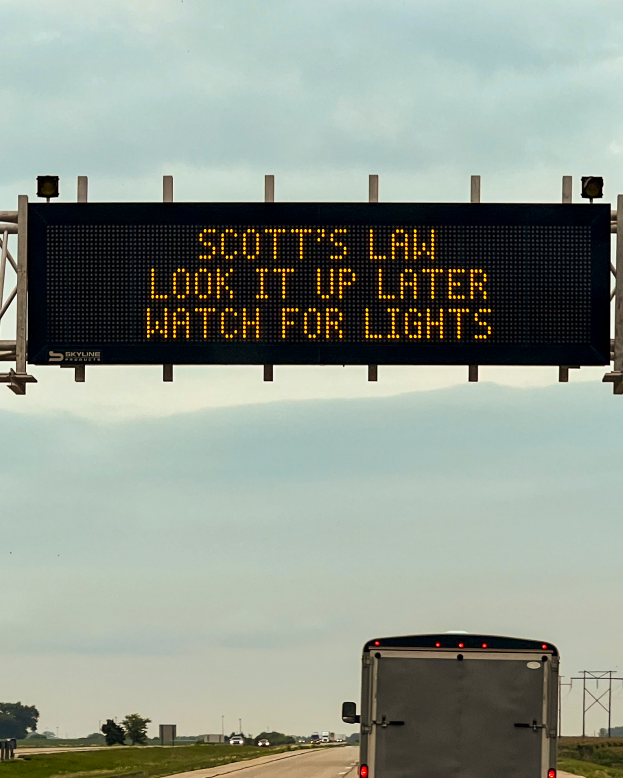 Ein Lkw fährt auf einer Autobahn mit einem Schild an der Seite, auf dem steht: "Scott's Law: Schau es später nach, achte auf Lichter", umgeben von Gras, Bäumen und Pfählen, mit einem klaren Himmel im Hintergrund.