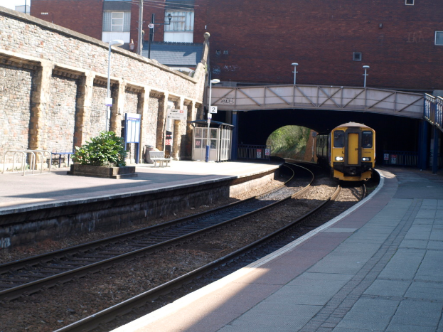 Ein Zug fährt auf Eisenbahnschienen mit Bahnsteigen auf beiden Seiten, eine Hauswand links, Straßenlaternen, Pflanzen, Bänke, eine Kabine, eine Brücke über die Bahnsteige und Gebäude und Masten im Hintergrund.