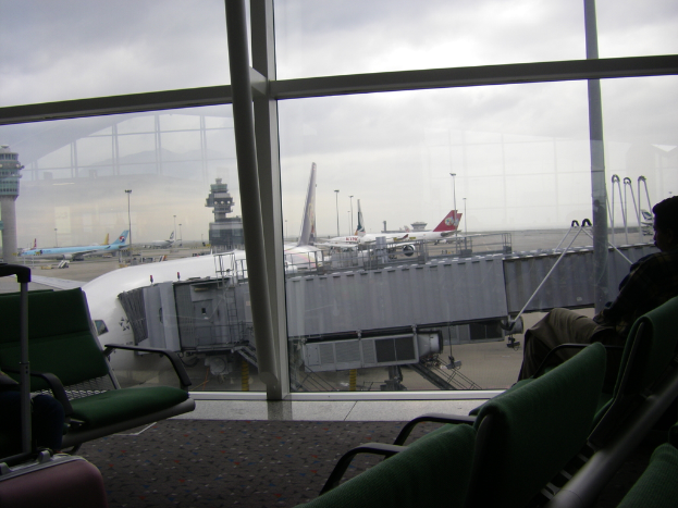 Ein Blick aus einem Fenster, der mehrere Stühle, einen Koffer auf der linken Seite, Flugzeuge und andere Abteile draußen zeigt.