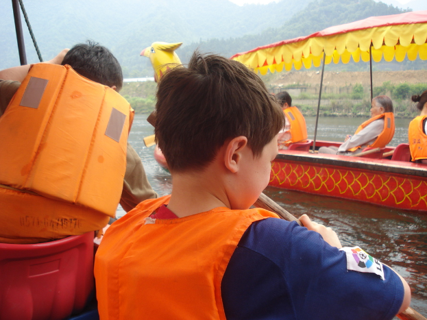 Eine friedliche Landschaft mit wenigen Menschen, Booten auf dem Wasser, Bäumen und Hügeln im Hintergrund.