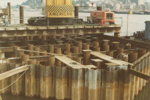 Baustelle mit einem Kran, der Stahlstangen von einem Haufen im Wasser hebt, umgeben von Gebäuden, Bäumen und einem klaren blauen Himmel.