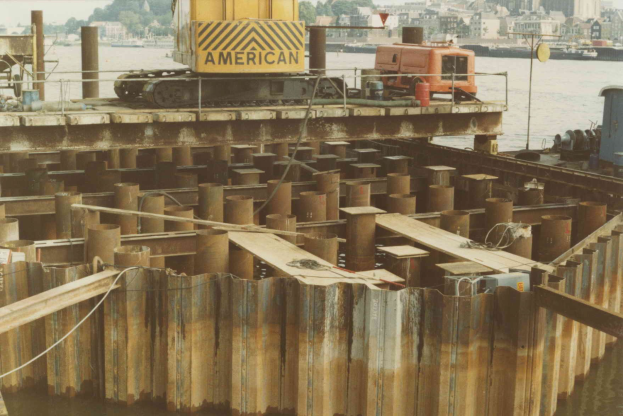 Baustelle mit einem Kran, der Stahlstangen von einem Haufen im Wasser hebt, umgeben von Gebäuden, Bäumen und einem klaren blauen Himmel.