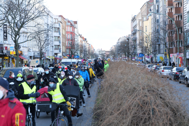 Eine große Gruppe von Menschen in Masken und Sicherheitswesten auf Fahrrädern auf einer von Bäumen gesäumten Straße mit Gebäuden, Laternenmasten, Texttafeln, Fahrzeugen, trockenem Gras und einem klaren blauen Himmel.