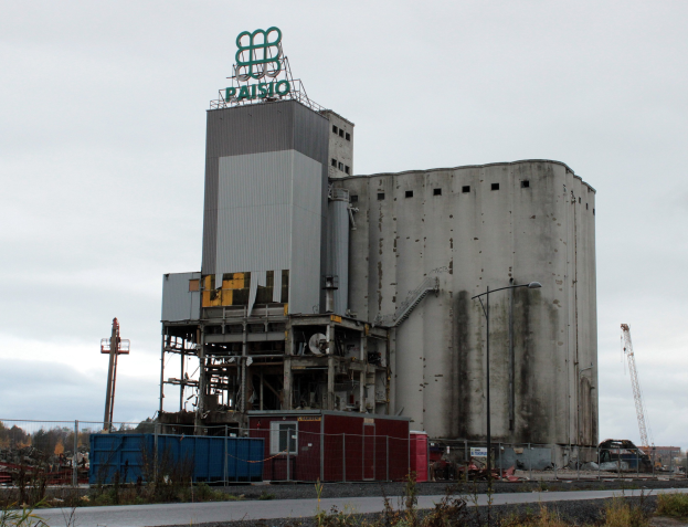 Großes Zementgebäude mit einem Zaun, Pfählen und Laternen, umgeben von Pflanzen, an der Seite einer Straße mit Bäumen und einem klaren blauen Himmel im Hintergrund.