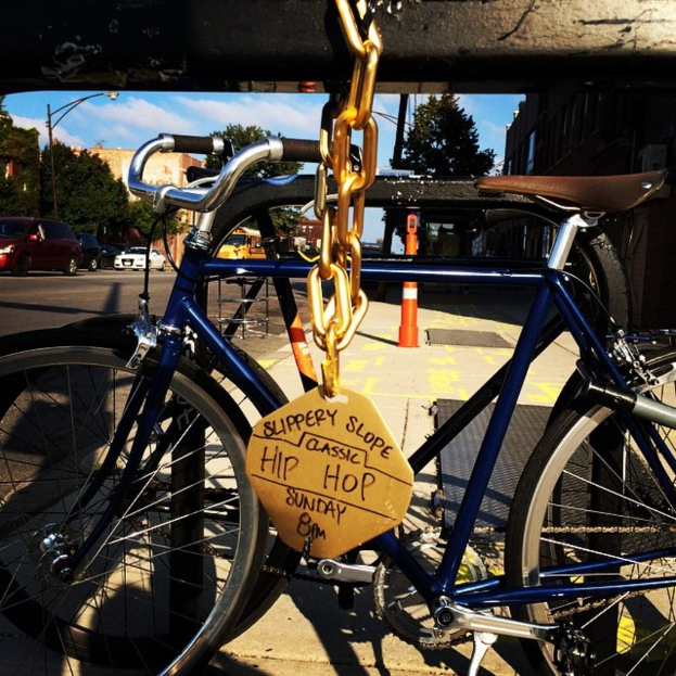 Eine Straße mit einem verriegelten Fahrrad auf dem Gehweg, Fahrzeuge im Hintergrund, Gebäude, Bäume, Pfosten, Gullis und ein bewölkter Himmel.