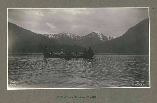 Schwarzes und weißes Foto einer Familie in einem Kanu auf einem See mit Bergen und Bäumen im Hintergrund, Himmel oben und Text unten.