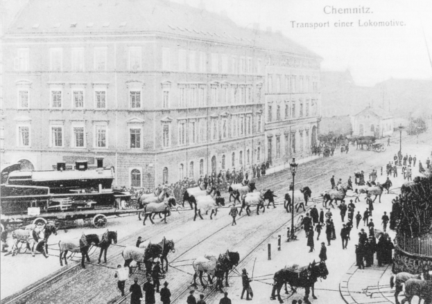 A black and white photo of a bustling city street with horse-drawn carriages, pedestrians, light poles, and buildings with windows, featuring text at the top.