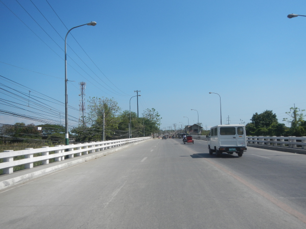 Ein weißer Lieferwagen fährt auf einer Autobahn mit Leitplanken, Laternenmasten, Strommasten mit Drähten, Bäumen und einem Turm im Hintergrund unter einem klaren blauen Himmel.