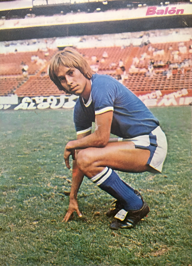 Ein Mann kniet auf einem Fußballfeld umgeben von sitzenden Personen mit Schildern und trägt Sportschuhe.