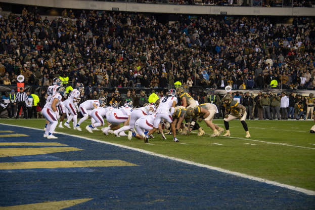 Ein Fußballspiel im Gange mit Spielern auf dem Boden in Sportbekleidung und Helmen, betrachtet von einem Stadion-Sitzbereich mit Zuschauern und Beschriftungen im Hintergrund.