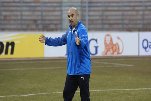 Ein Mann in einer blauen Jacke steht auf einem Fussballfeld, umgeben von Tafeln mit Text und Logos, entschlossen.