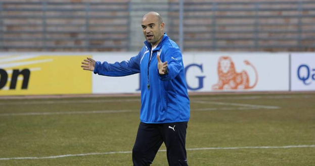 Ein Mann in einer blauen Jacke steht auf einem Fussballfeld, umgeben von Tafeln mit Text und Logos, entschlossen.