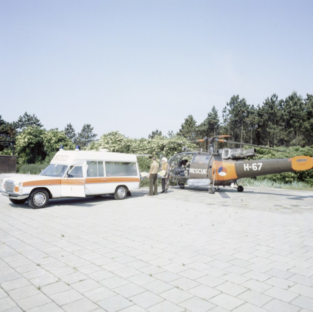 Ein Krankenwagen neben einem Hubschrauber auf einem gefliesten Boden geparkt, mit ein paar Menschen in der N├Ąhe, B├Ąumen und einem klaren blauen Himmel im Hintergrund.