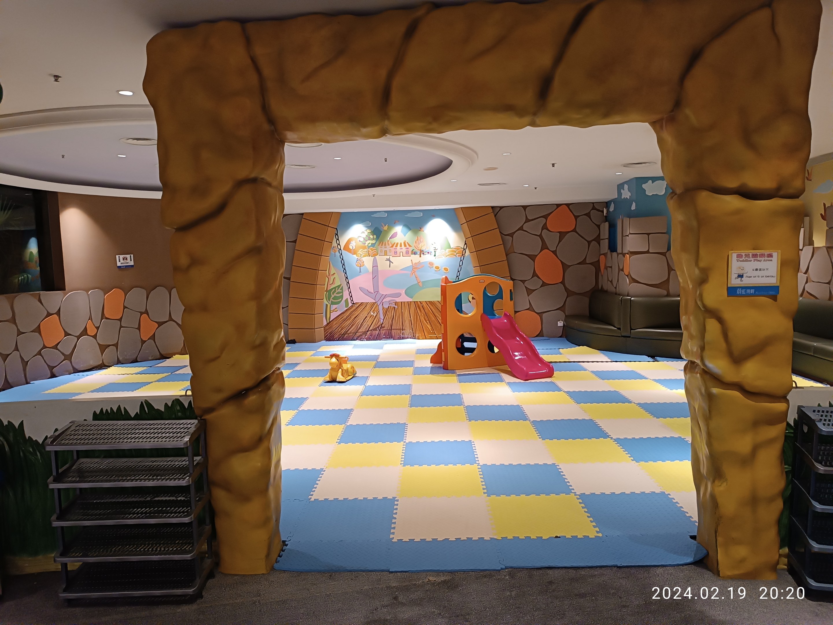 Kinderspielbereich mit einem zentralen Torbogen, Spielzeug auf dem Boden, Regalen an beiden Seiten, einer bemalten Wand im Hintergrund und Deckenbeleuchtung.