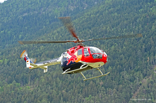 Roter Bullen-Hubschrauber fliegt über einen grünen Wald mit Bäumen auf Hügeln unter einem klaren blauen Himmel.
