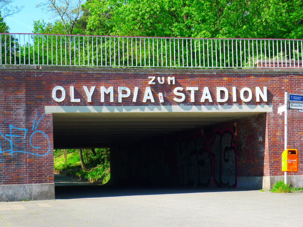 Der Eingang zum Olympiastadion in Berlin, Deutschland, mit einer Brücke, einem Metallzaun, einem Schild, einer Box, Pflanzen, Gras, einer Gruppe von Bäumen und einem bewölkten Himmel.