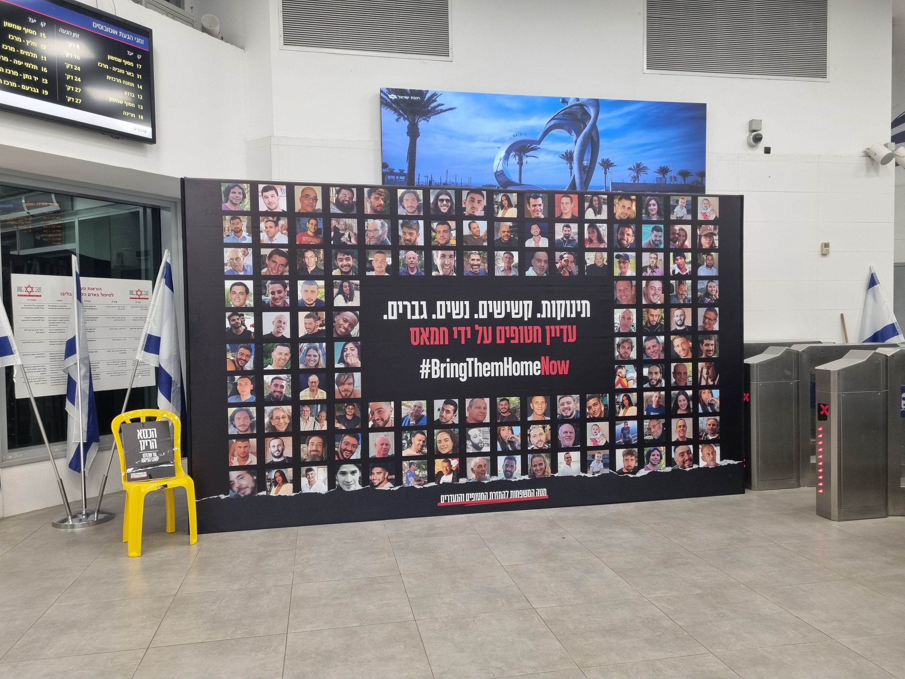 Eine große Wand zeigt zahlreiche Fotos, einen gelben Stuhl, israelische Regierungsflaggen auf Stangen, eine Tafel mit der Aufschrift "Bring Them Home Now", einen wandmontierten Bildschirm, Deckenleuchten und andere Gegenstände.