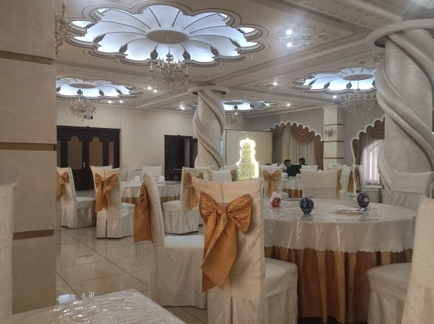 Ein großer Festsaal mit Tischen und Stühlen, die mit Tüchern drapiert und mit verschiedenen Gegenständen geschmückt sind, mit Türen und Vorhängen an den Wänden und von Deckenleuchten und Kronleuchtern beleuchtet.