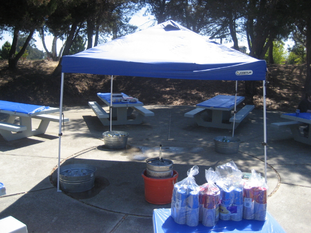 Party cups in covers with price tags on a table, buckets with objects under a tent, benches with objects around the tent, trees and a wooden house in the background.