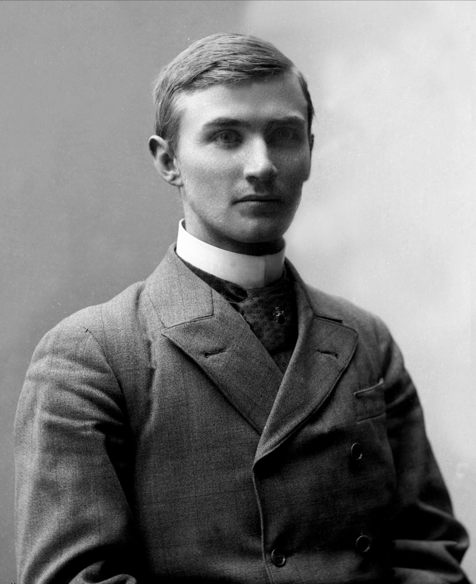 Schwarzes und weißes Porträt eines Mannes in Anzug und Krawatte, der mit gefalteten Händen und ernstem Gesichtsausdruck steht.