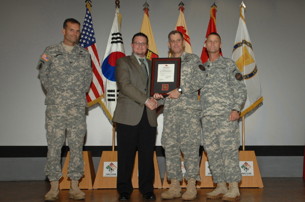 Vier Männer auf einer Bühne: drei in Armeeuniformen und einer in Blazer, Krawatte und Hemd, die Hände schütteln und einen Preis überreichen, mit sechs Fahnen an Stangen und einer weißen Wand im Hintergrund.