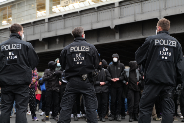 Eine Gruppe uniformierter Polizisten steht vor einer Menge von Menschen in schwarzen Uniformen und Masken, mit einer Brücke und einem Gebäude im Hintergrund, während einer Protestaktion in einer Stadt.