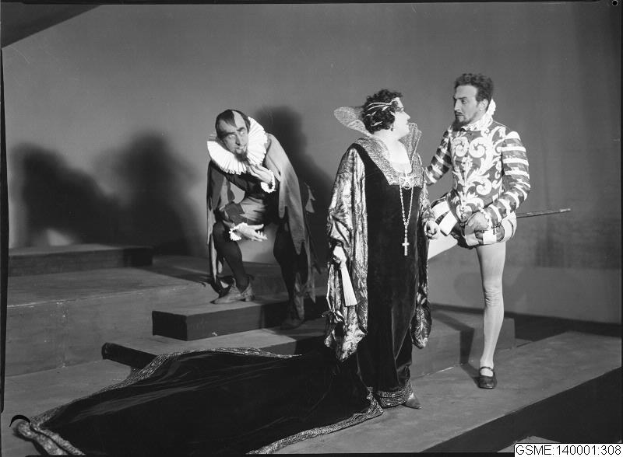 Schwarzes und weißes Foto von drei Menschen auf einer Bühne, wobei einer ein Kostüm trägt und einen Stock hält, ein anderer auf einer Treppe sitzt und unten ein Text mit "The Phantom of the Opera" steht.