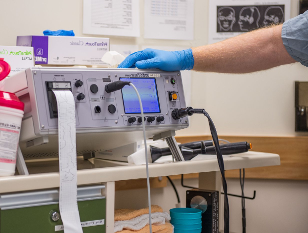Eine Person in blauen Handschuhen arbeitet an einem medizinischen Gerät an einem unordentlichen Tisch in einem Krankenhaussetting, mit Papieren an der Wand im Hintergrund.