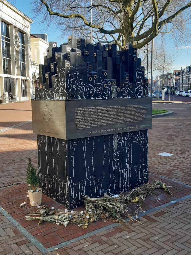 Denkmal für Holocaust-Opfer in einem Stadtplatz mit einem Baum, Topfpflanze, verstreuten trockenen Blättern und umliegenden städtischen Elementen.
