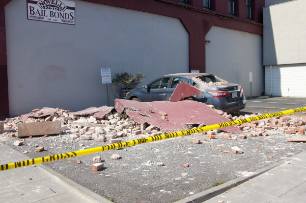 Eine zerbrochene Ziegelwand liegt auf dem Boden mit verstreuten roten Ziegeln, ein geparktes Auto, ein Topf mit einer Pflanze und ein Gebäude mit einer 'Kautionsbürgschaft'-Plakette im Hintergrund.