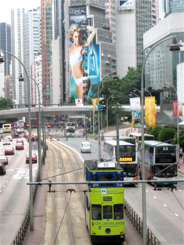 Eine belebte Stadtstraße mit verschiedenen Fahrzeugen, darunter Doppeldeckerbusse, Autos und andere Fahrzeuge, unter einem klaren blauen Himmel, mit hohen Gebäuden, einer Brücke und verschiedenen Straßenmöbeln.