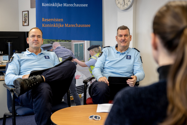 Zwei Polizeibeamte sitzen an einem Tisch mit einer Frau, umgeben von Papieren und Gegenständen, mit einem Banner, einer Uhr und Fotorahmen an der Wand und einem weiteren Tisch mit einem Monitor und Tastatur links daneben.