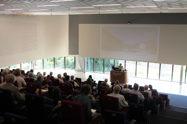 Ein Seminarsaal an einer Universität mit vielen Menschen, die aufmerksam einer Präsentation an der Front zugewandt sind, wo eine Person an einem großen Tisch die Präsentation erklärt, mit Glasfenstern im Hintergrund, die Bäume und Gebäude zeigen.